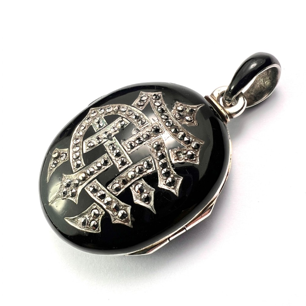 An oval Victorian silver locket rendered in black enamel with an intricate marcasite-set AEI monogram on the front surface.