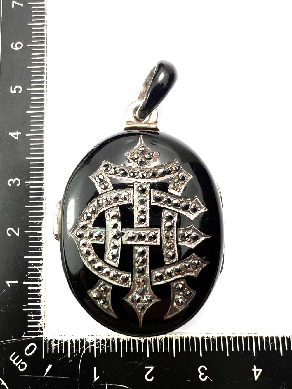 An oval Victorian silver locket rendered in black enamel with an intricate marcasite-set AEI monogram on the front surface. View IX