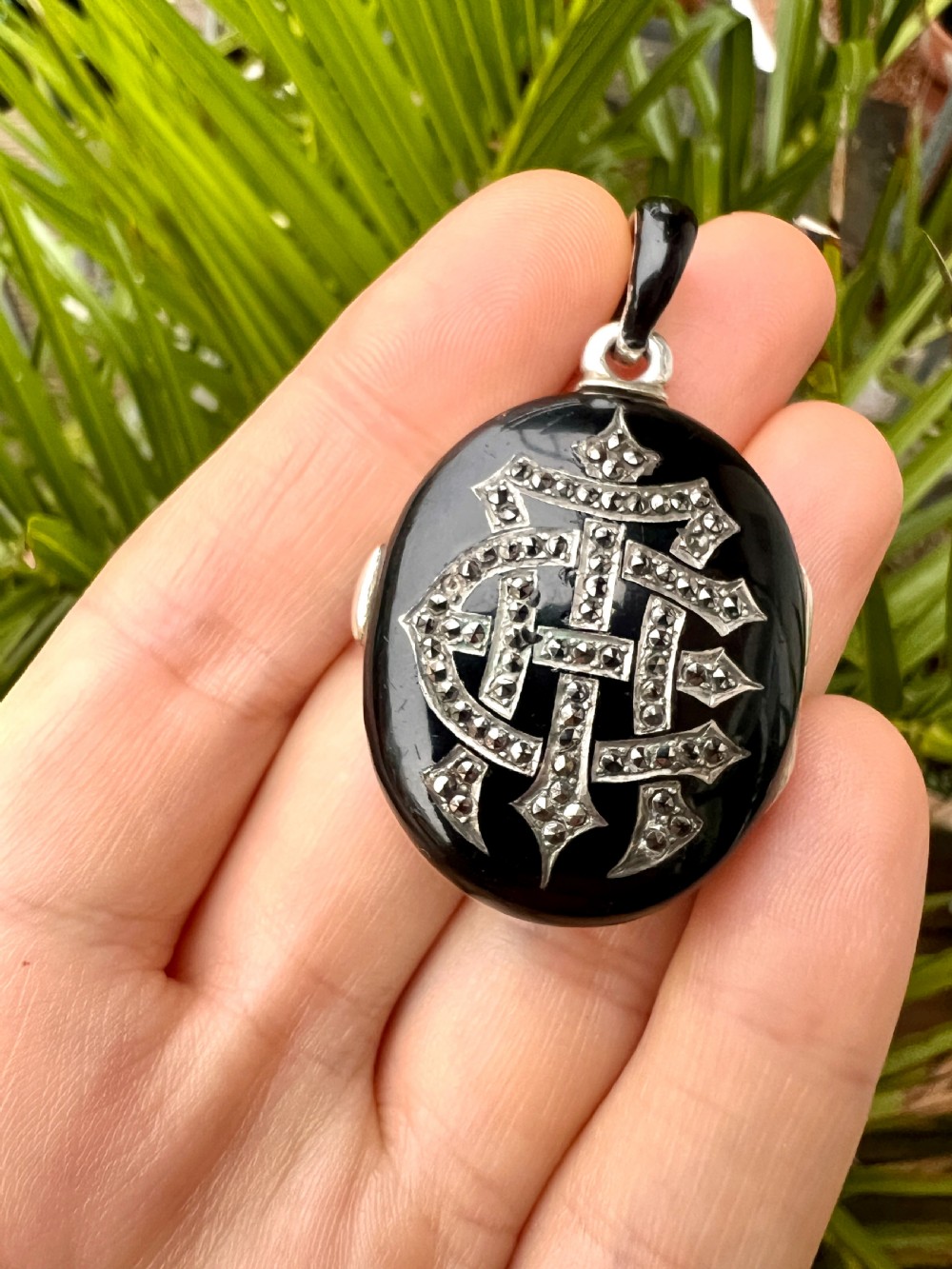 An oval Victorian silver locket rendered in black enamel with an intricate marcasite-set AEI monogram on the front surface. View X