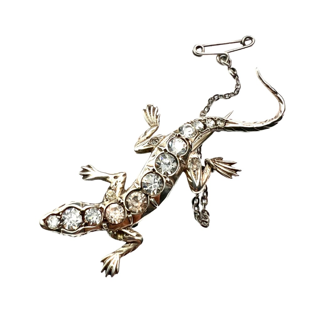 An Art Deco silver brooch shaped like a lizard, encrusted with round clear paste stones along its spine and head, featuring an attached safety chain.
