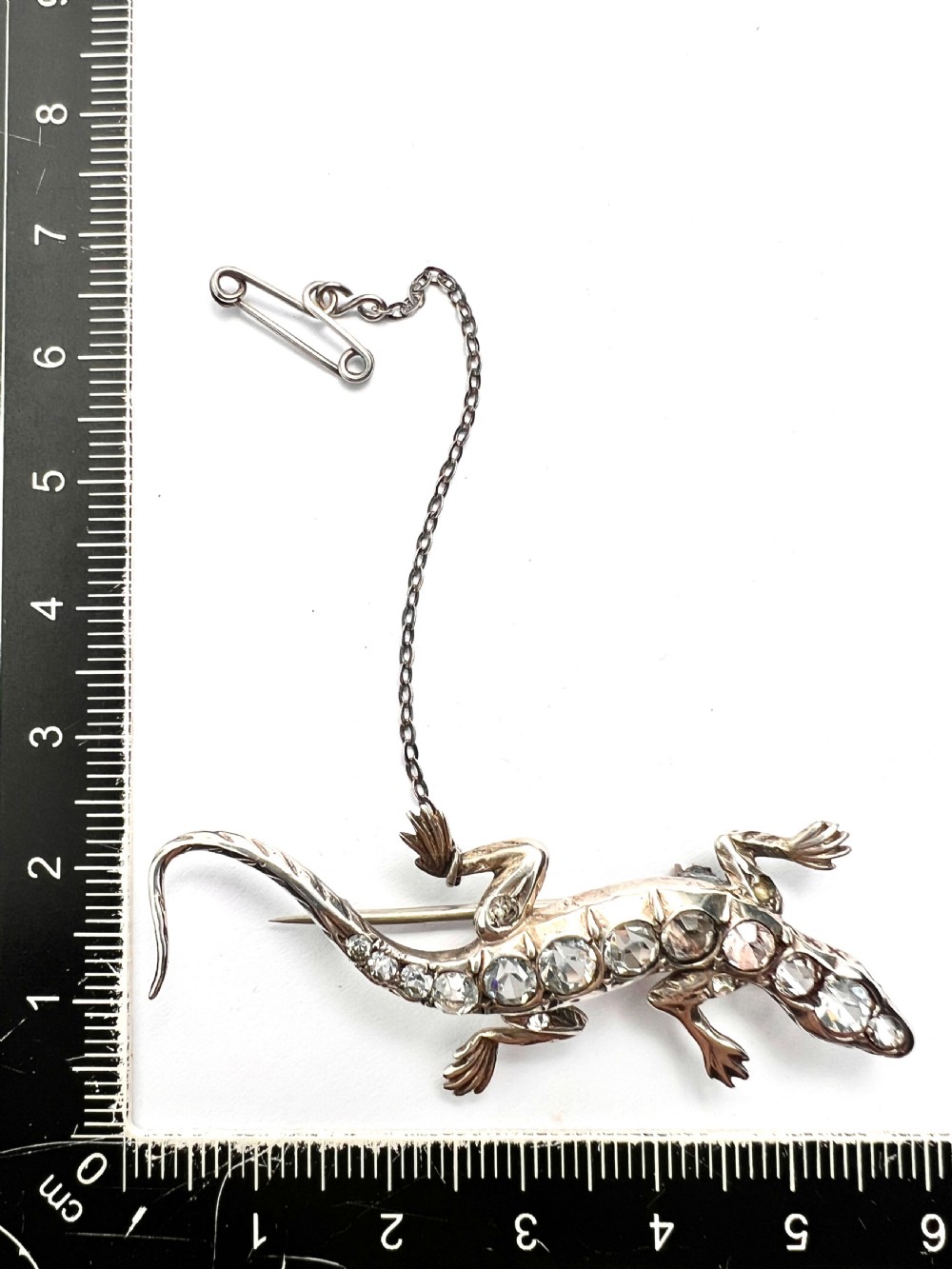 An Art Deco silver brooch shaped like a lizard, encrusted with round clear paste stones along its spine and head, featuring an attached safety chain. View VII