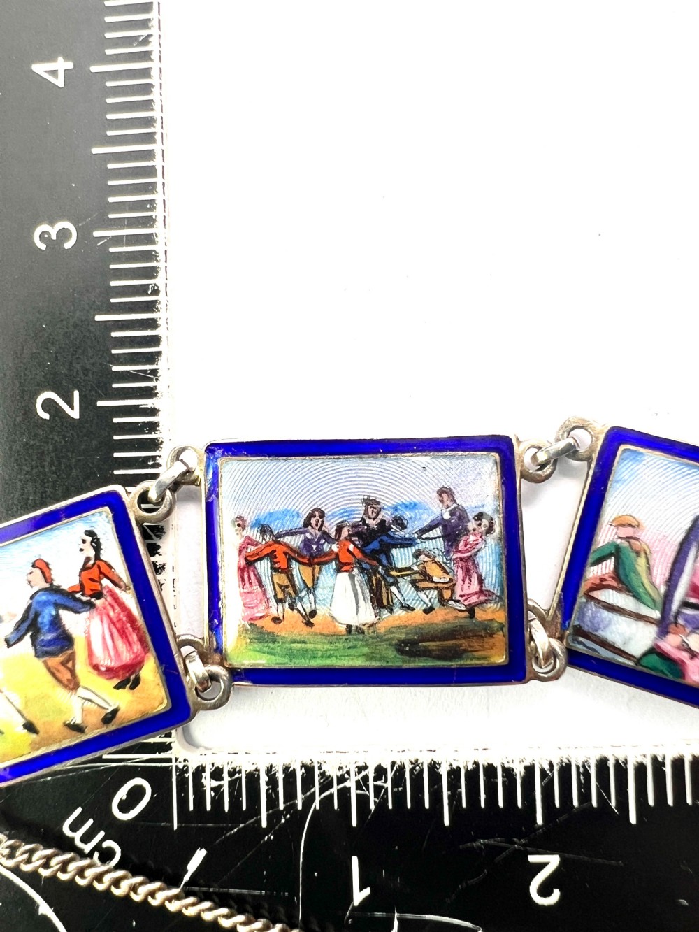 A vintage silver link bracelet featuring rectangular panels decorated with hand-painted miniature genre scenes bordered in cobalt blue enamel. View V