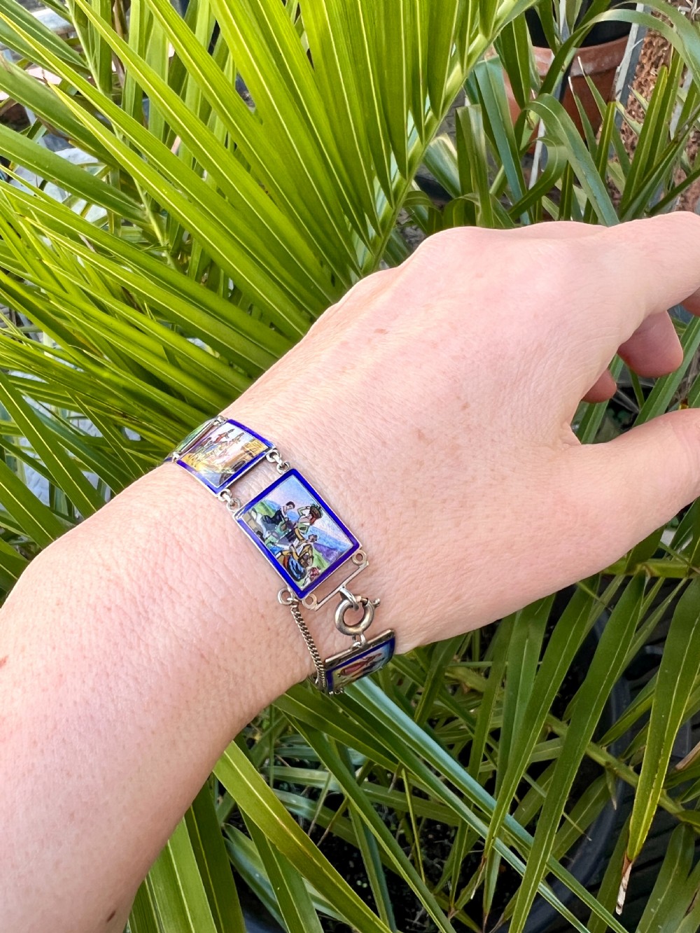 A vintage silver link bracelet featuring rectangular panels decorated with hand-painted miniature genre scenes bordered in cobalt blue enamel. View VI