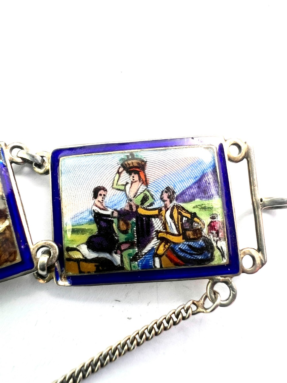 A vintage silver link bracelet featuring rectangular panels decorated with hand-painted miniature genre scenes bordered in cobalt blue enamel. View X