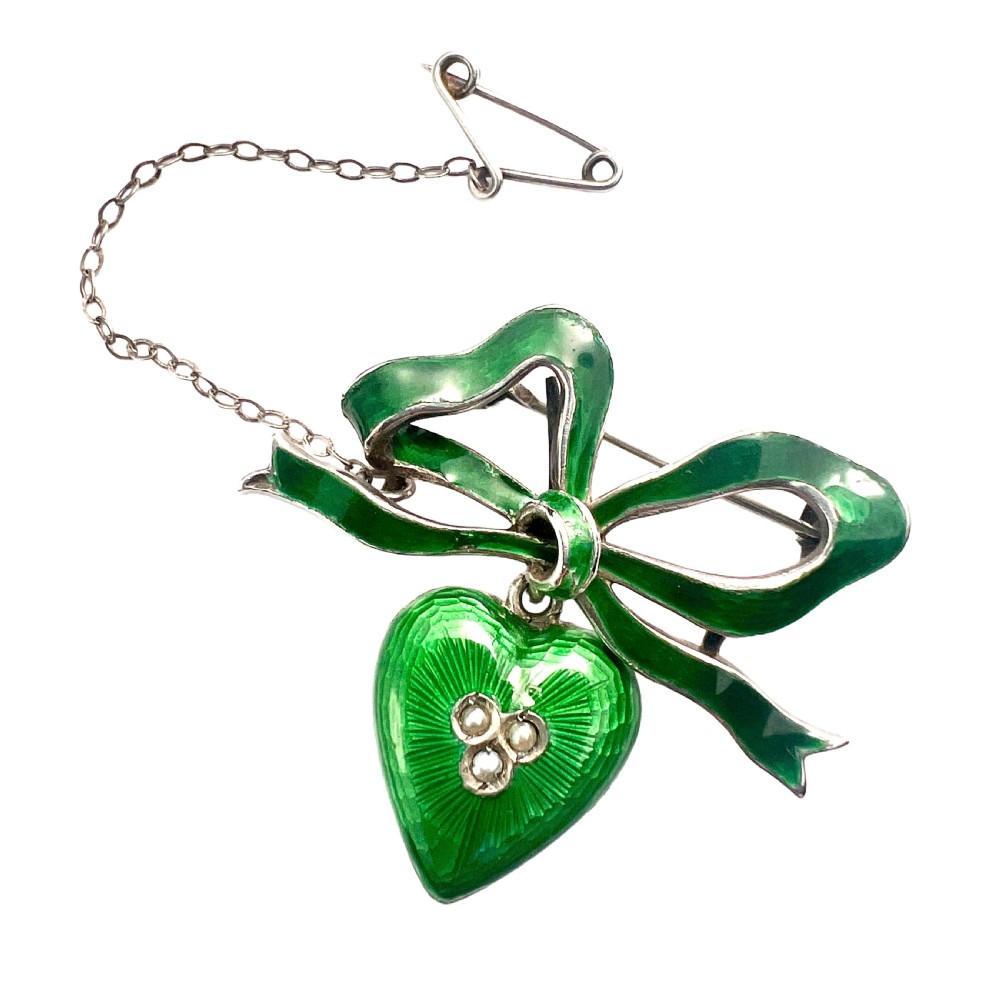 An antique silver brooch featuring a green translucent enamel ribbon bow with a hanging green enamel heart drop set with three small seed pearls.