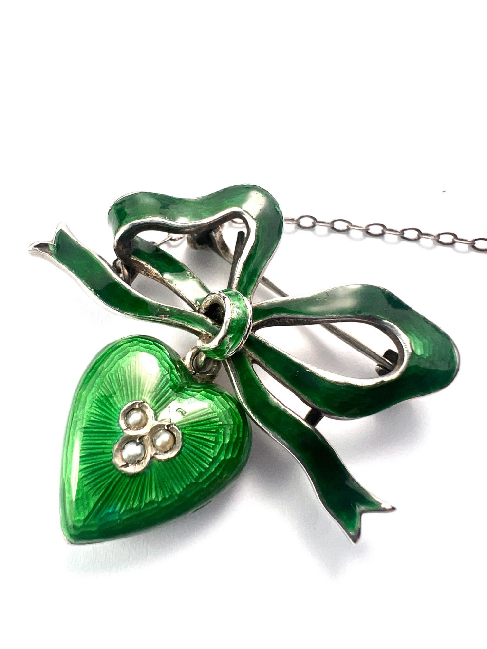 An antique silver brooch featuring a green translucent enamel ribbon bow with a hanging green enamel heart drop set with three small seed pearls. View V
