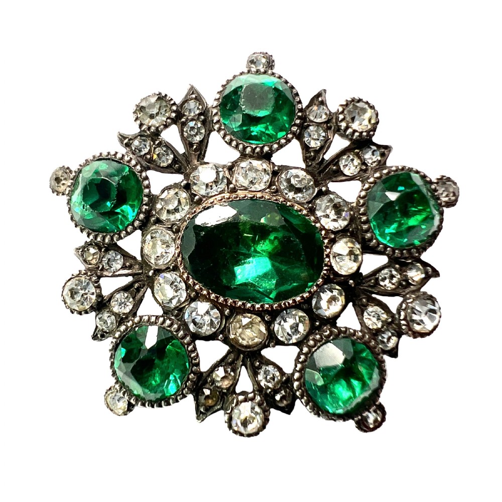 A silver cluster brooch featuring an oval green paste stone at the centre, encircled by smaller clear stones and outer green paste stones.