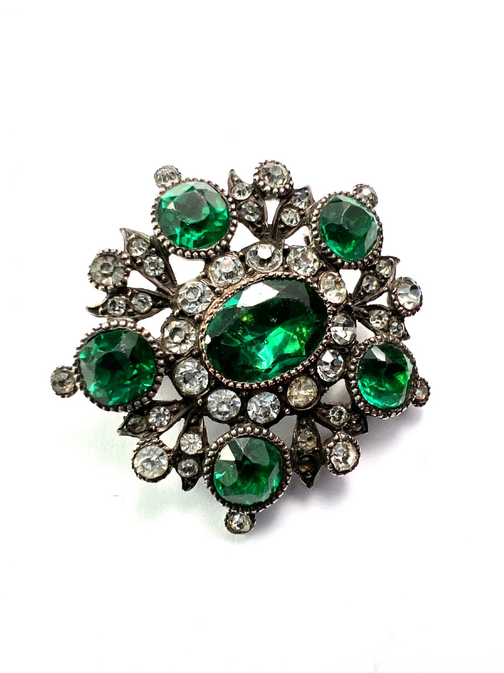 A silver cluster brooch featuring an oval green paste stone at the centre, encircled by smaller clear stones and outer green paste stones. View II