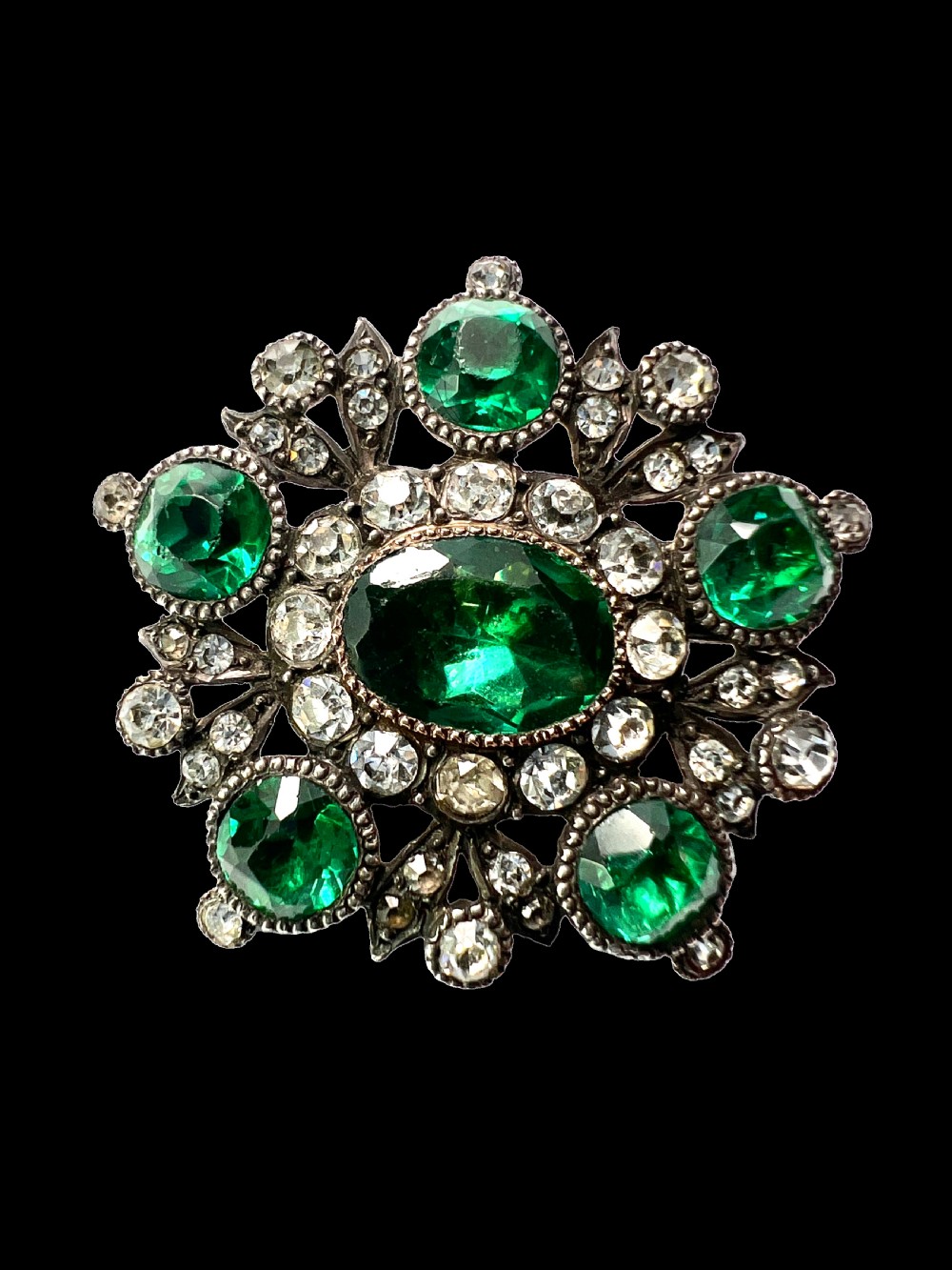 A silver cluster brooch featuring an oval green paste stone at the centre, encircled by smaller clear stones and outer green paste stones. View III
