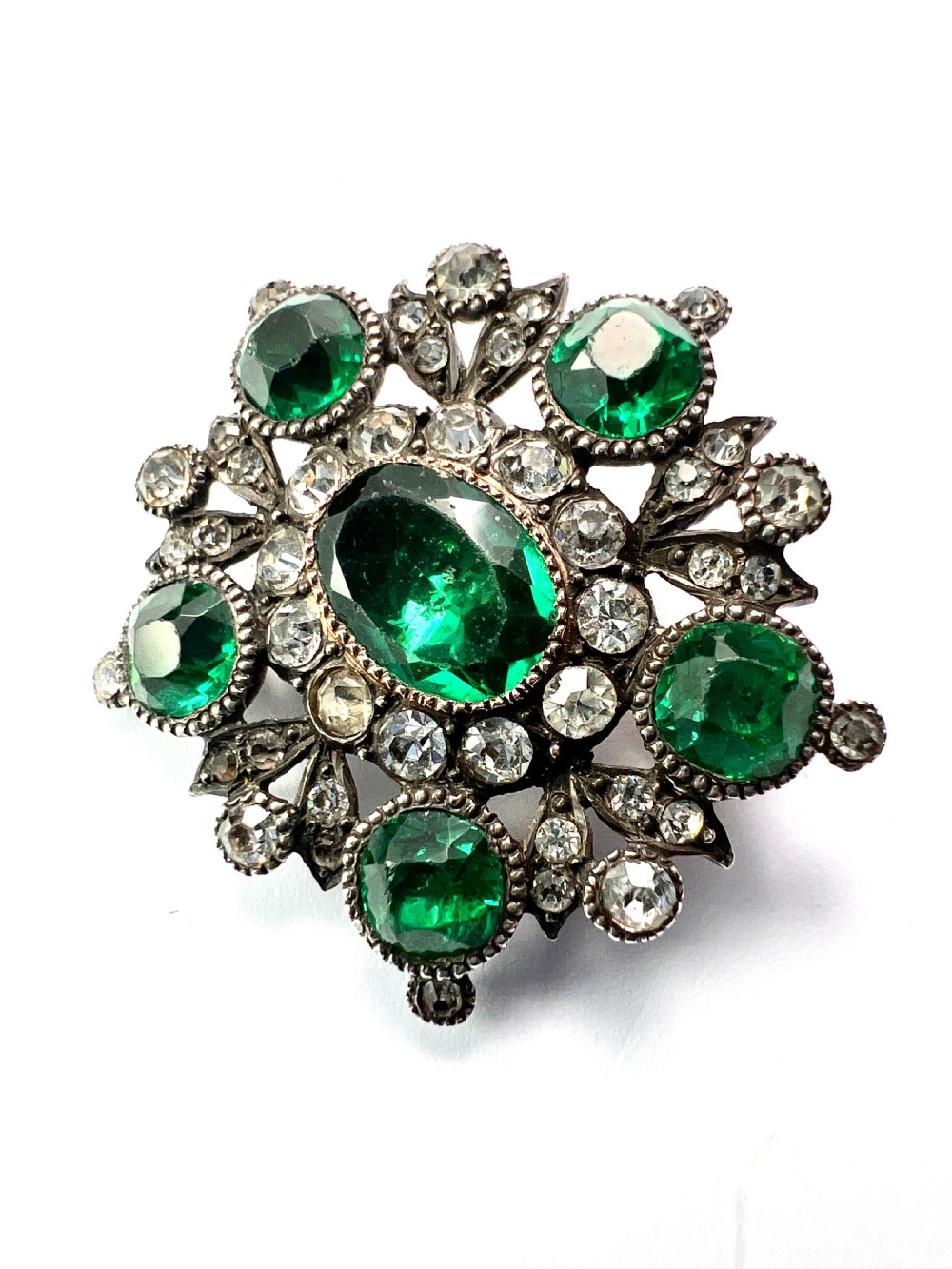 A silver cluster brooch featuring an oval green paste stone at the centre, encircled by smaller clear stones and outer green paste stones. View IV