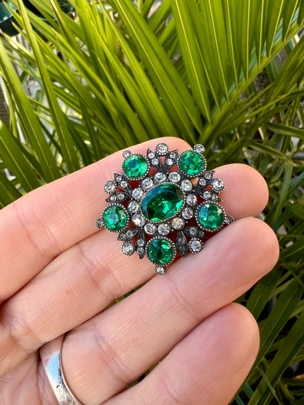 A silver cluster brooch featuring an oval green paste stone at the centre, encircled by smaller clear stones and outer green paste stones. View VII