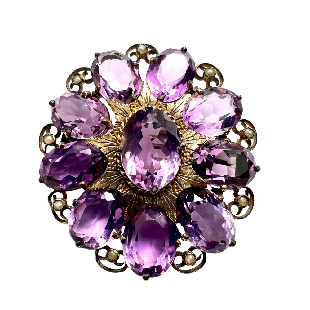A circular Victorian gold brooch featuring a cluster of oval faceted amethysts around a central oval amethyst, bordered by textured gold leaf details and scrollwork set with seed pearls.