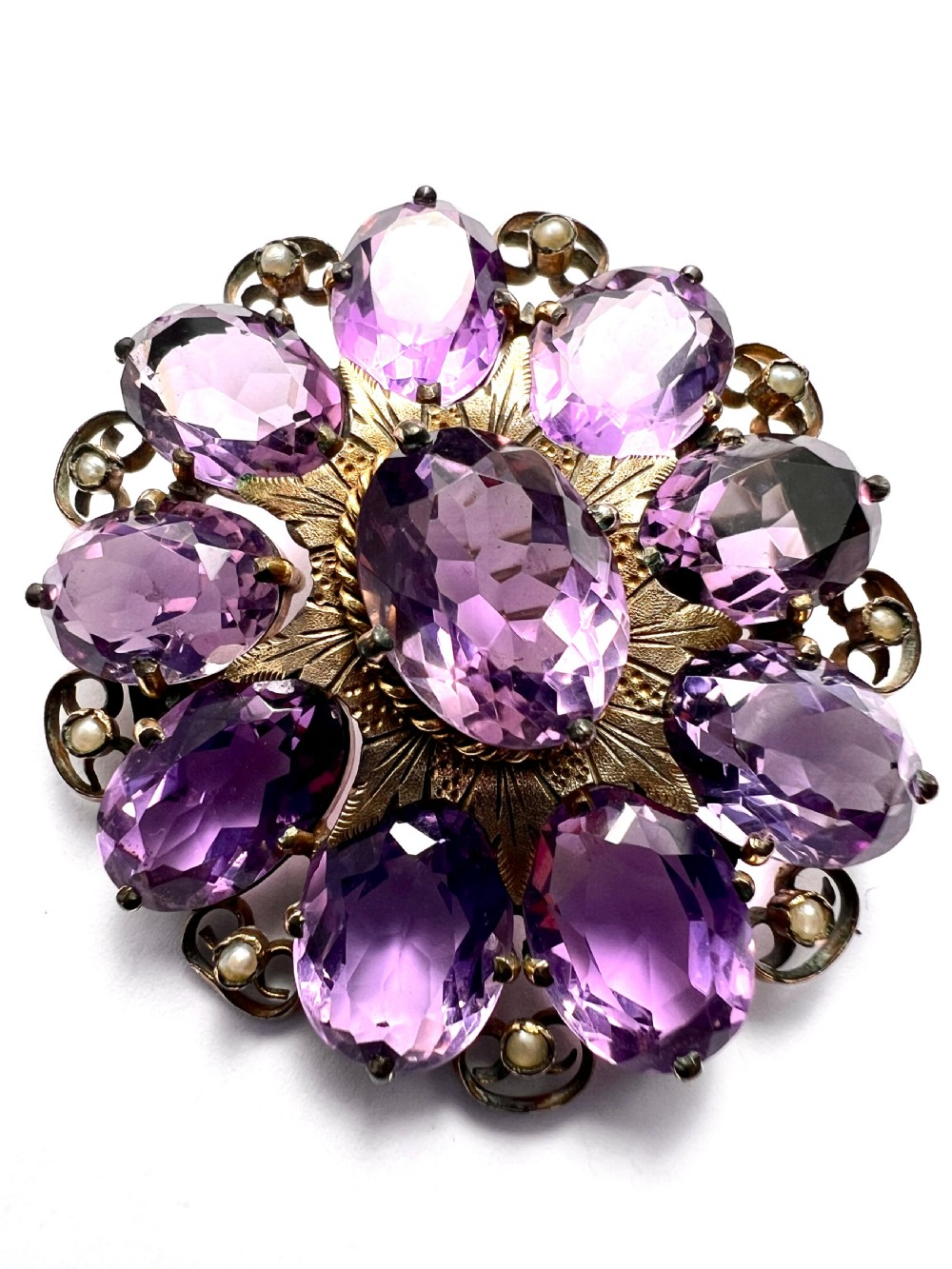 A circular Victorian gold brooch featuring a cluster of oval faceted amethysts around a central oval amethyst, bordered by textured gold leaf details and scrollwork set with seed pearls. View II