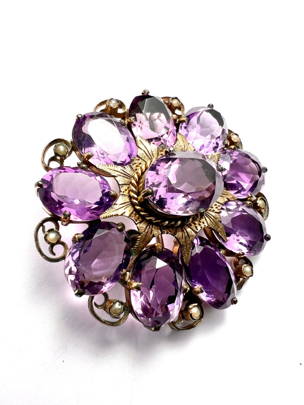 A circular Victorian gold brooch featuring a cluster of oval faceted amethysts around a central oval amethyst, bordered by textured gold leaf details and scrollwork set with seed pearls. View III