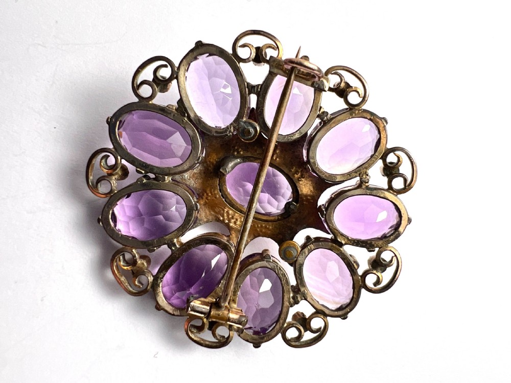 A circular Victorian gold brooch featuring a cluster of oval faceted amethysts around a central oval amethyst, bordered by textured gold leaf details and scrollwork set with seed pearls. View IV