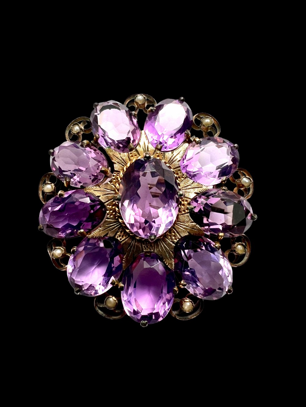 A circular Victorian gold brooch featuring a cluster of oval faceted amethysts around a central oval amethyst, bordered by textured gold leaf details and scrollwork set with seed pearls. View V