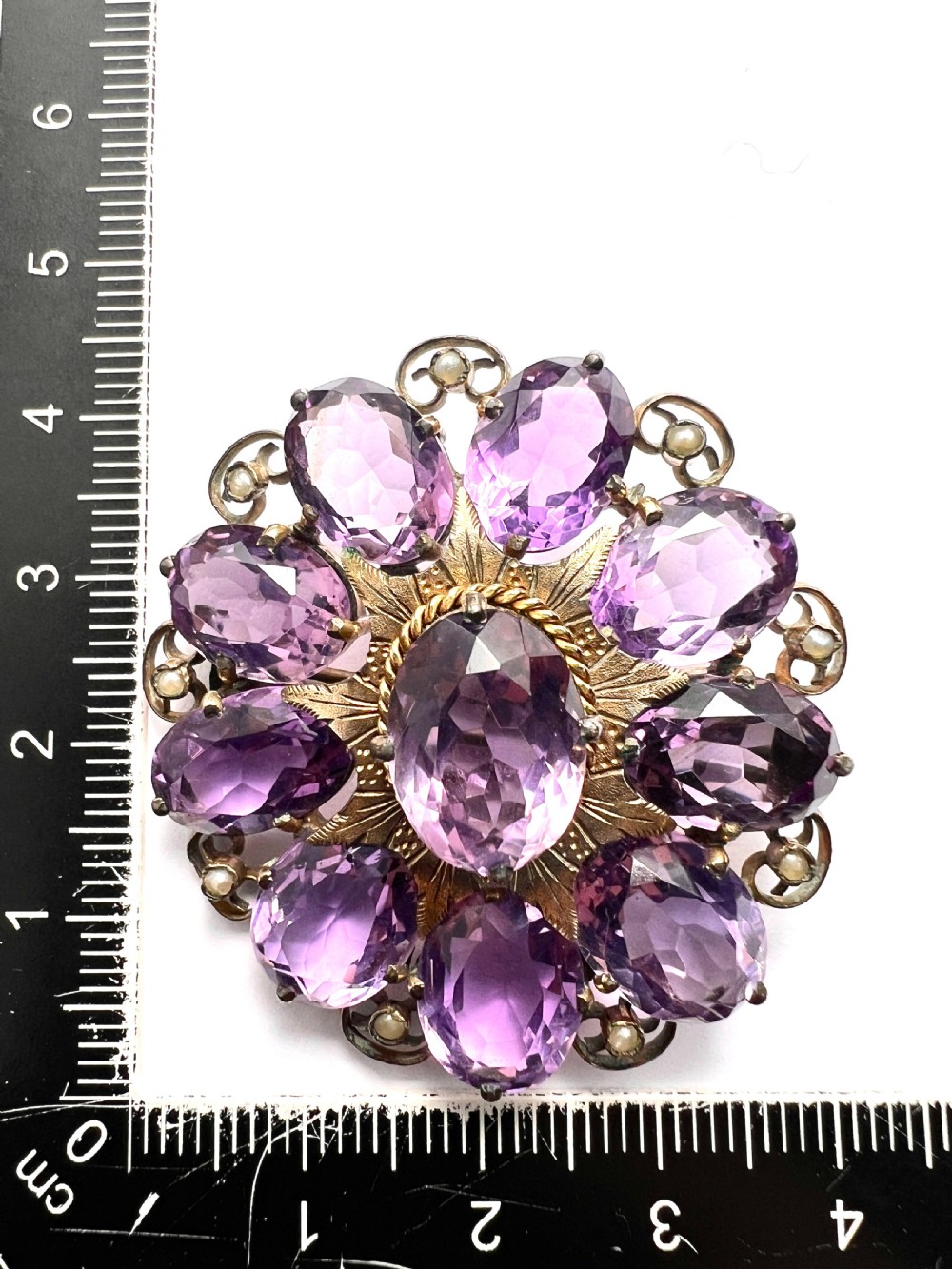 A circular Victorian gold brooch featuring a cluster of oval faceted amethysts around a central oval amethyst, bordered by textured gold leaf details and scrollwork set with seed pearls. View VI