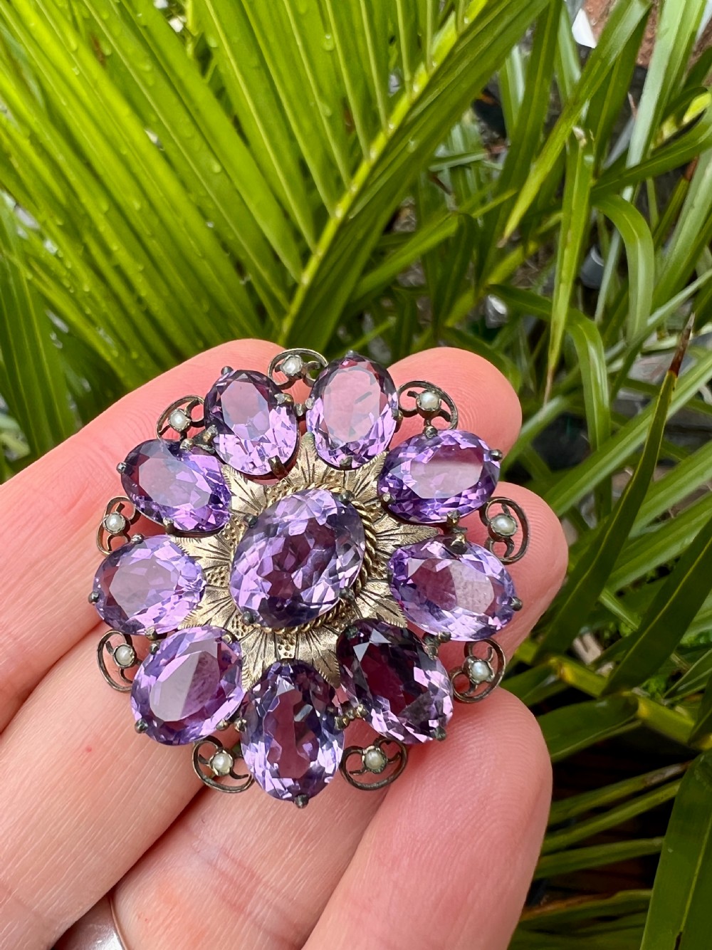 A circular Victorian gold brooch featuring a cluster of oval faceted amethysts around a central oval amethyst, bordered by textured gold leaf details and scrollwork set with seed pearls. View VII