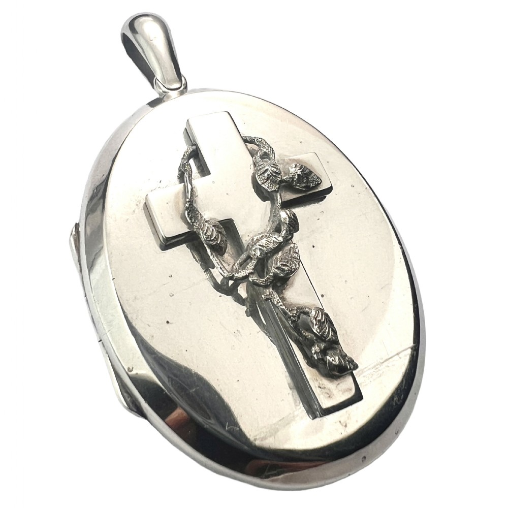 A large oval silver locket featuring a central raised crucifix motif draped with sculpted ivy foliage, fitted with an oval bail at the top.