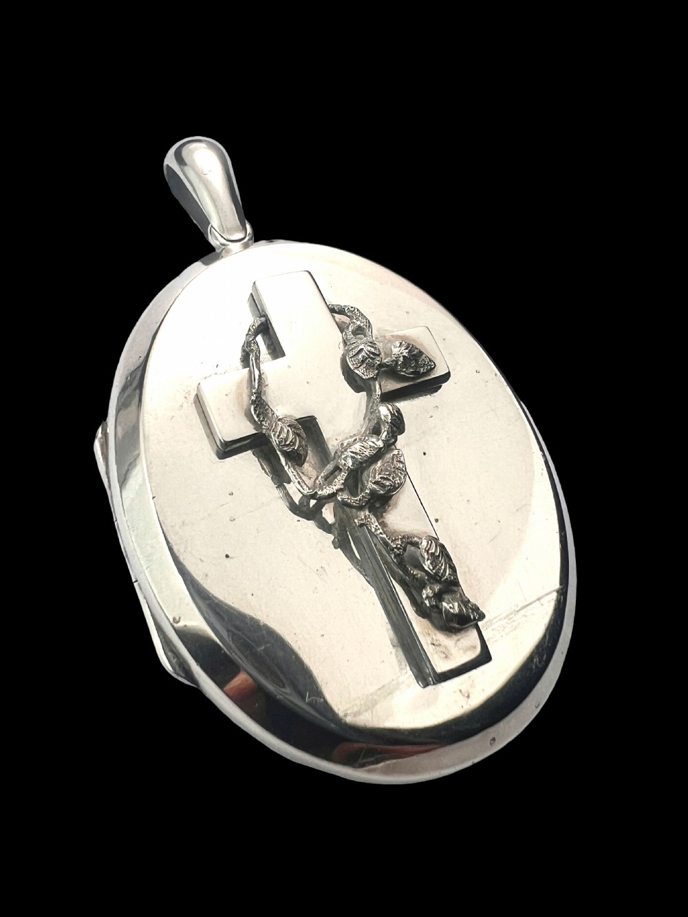 A large oval silver locket featuring a central raised crucifix motif draped with sculpted ivy foliage, fitted with an oval bail at the top. View VI