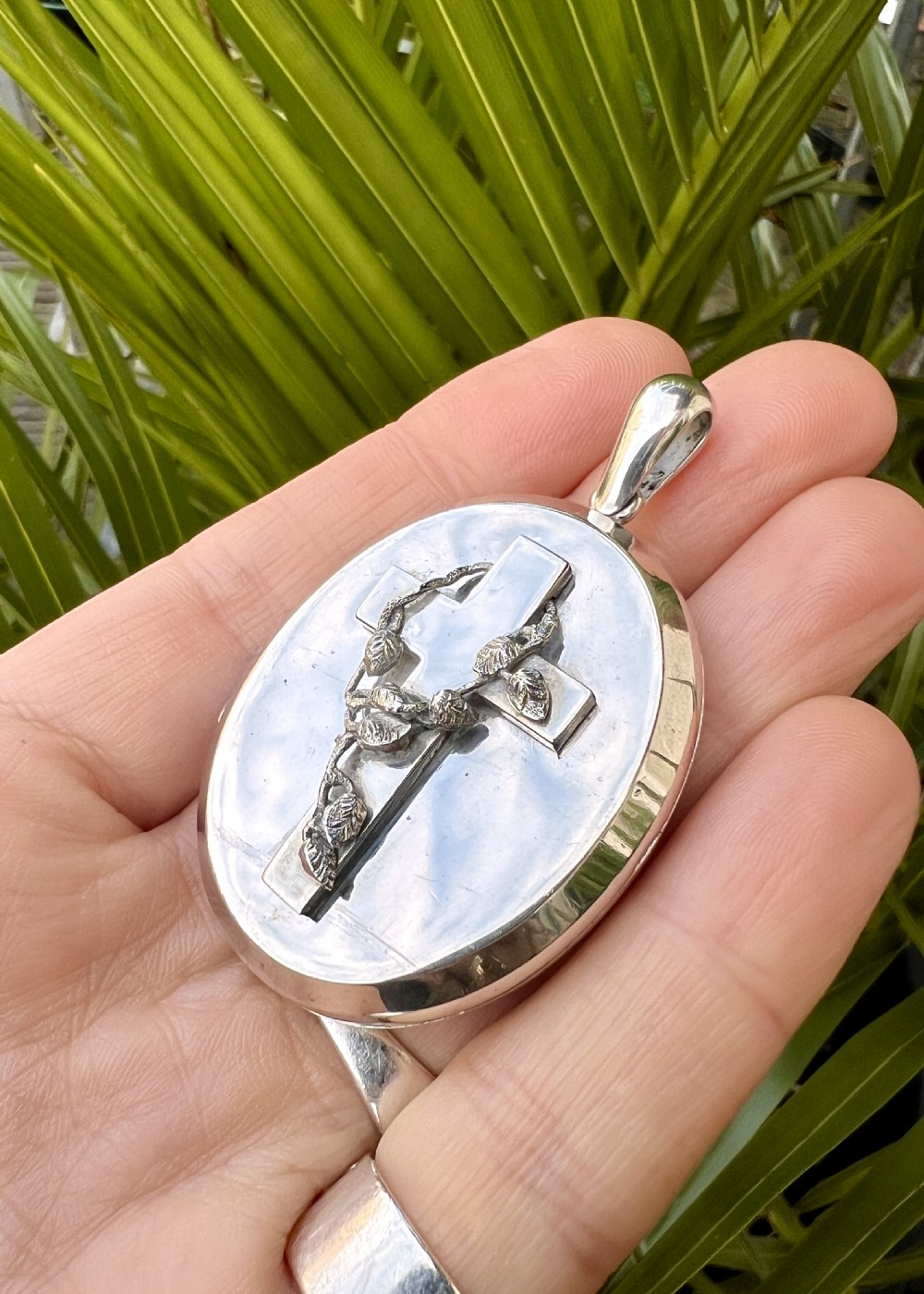 A large oval silver locket featuring a central raised crucifix motif draped with sculpted ivy foliage, fitted with an oval bail at the top. View VIII