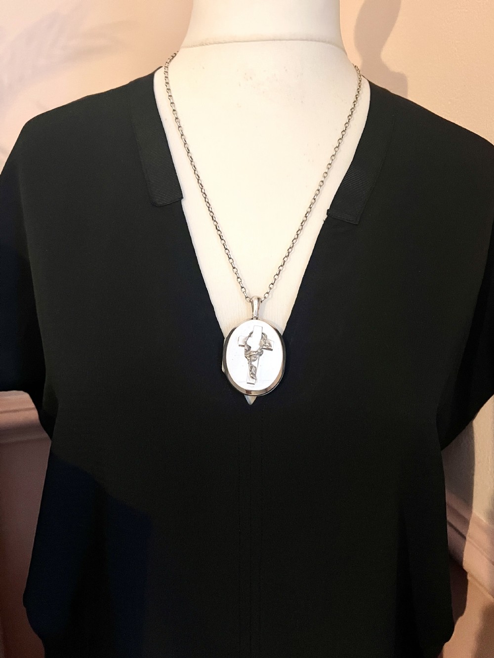 A large oval silver locket featuring a central raised crucifix motif draped with sculpted ivy foliage, fitted with an oval bail at the top. View IX