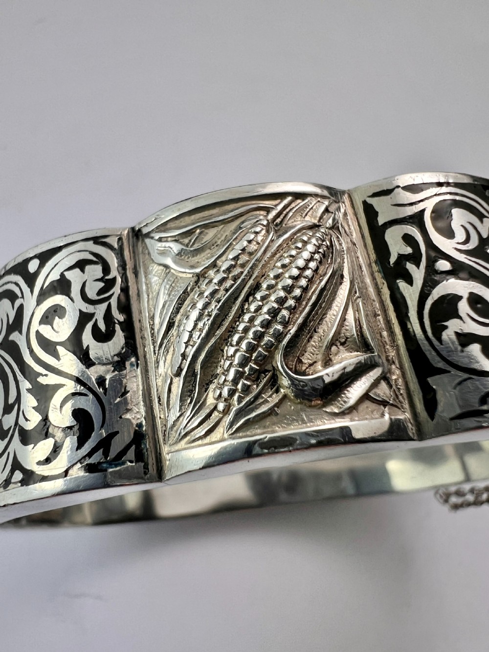A vintage Portuguese silver hinged cuff bangle featuring engraved foliate scrollwork and corn motifs with dark niello detailing and a safety chain. View IX