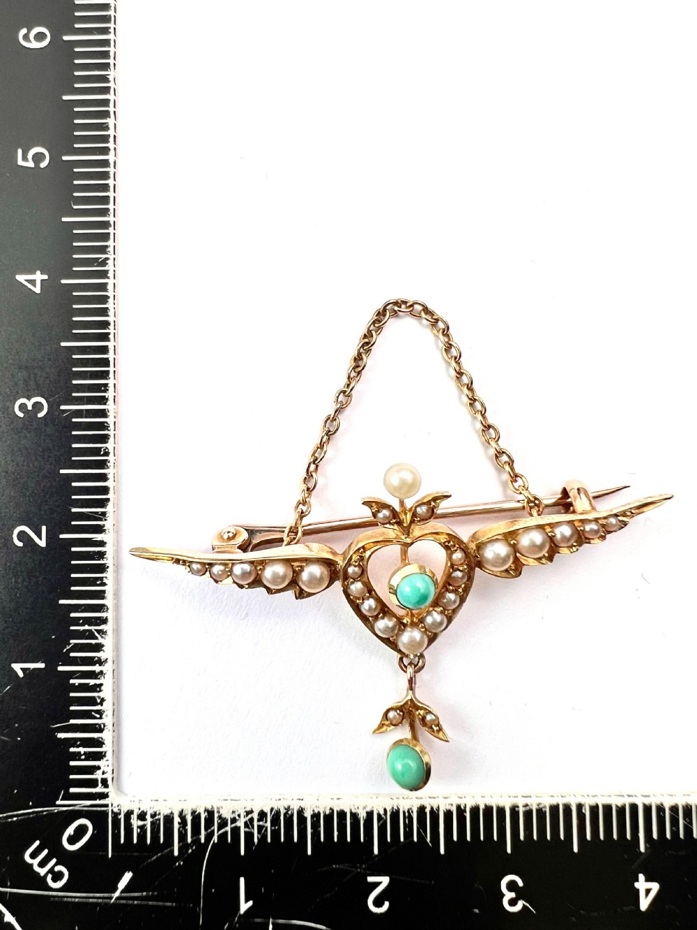 An Edwardian 15ct gold brooch featuring a winged heart motif set with seed pearls and turquoise cabochons with a draped lower chain. View II