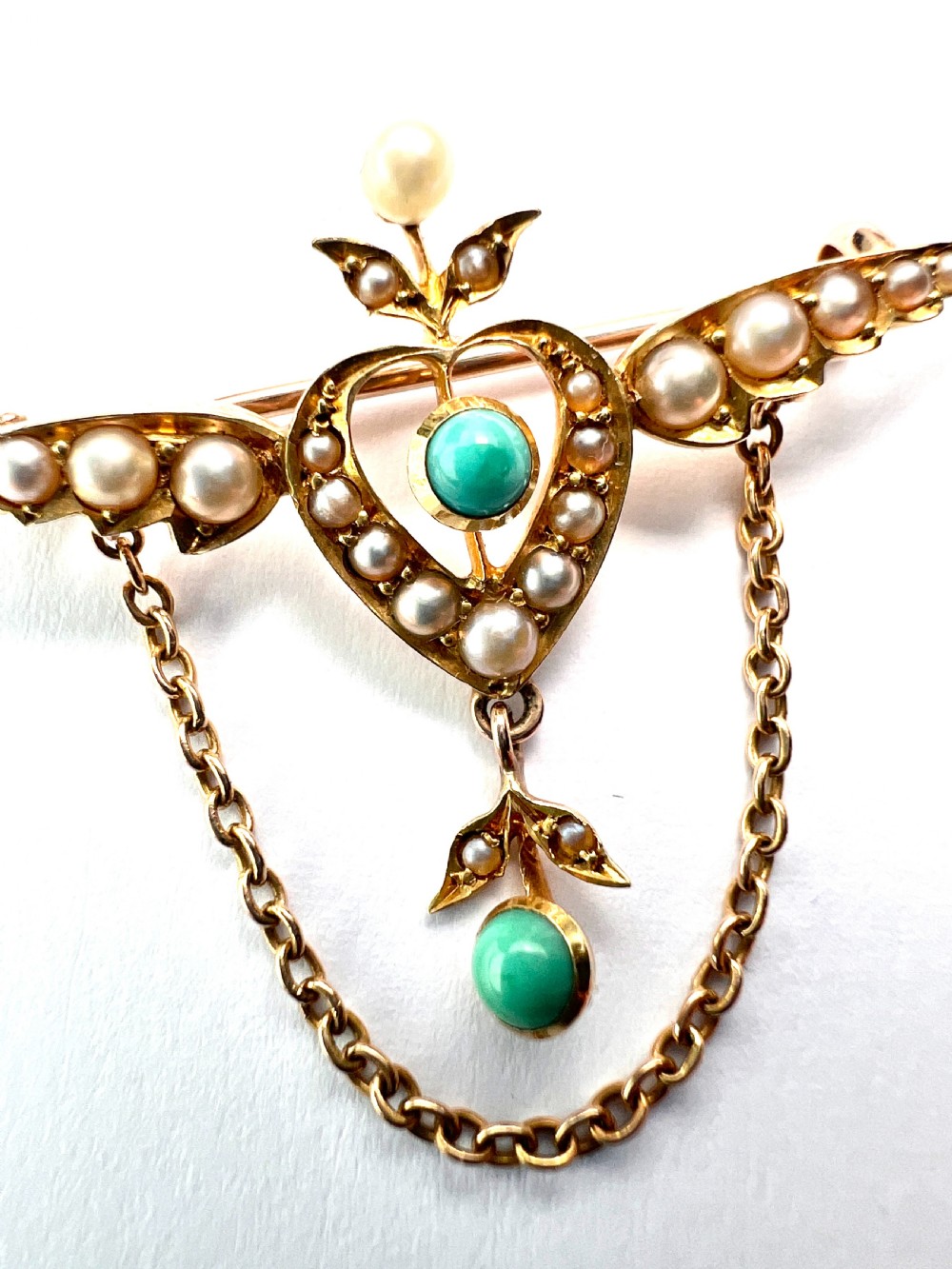 An Edwardian 15ct gold brooch featuring a winged heart motif set with seed pearls and turquoise cabochons with a draped lower chain. View IX
