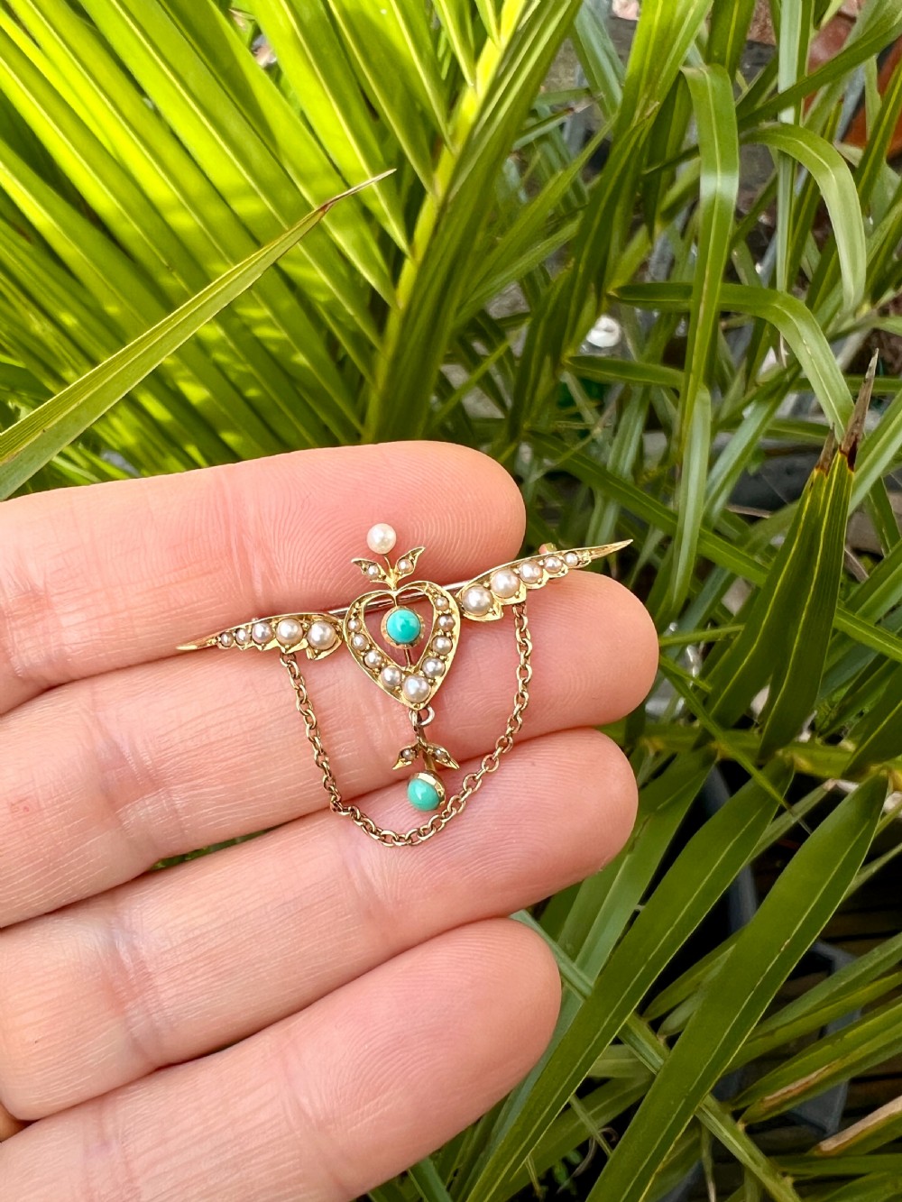 An Edwardian 15ct gold brooch featuring a winged heart motif set with seed pearls and turquoise cabochons with a draped lower chain. View X