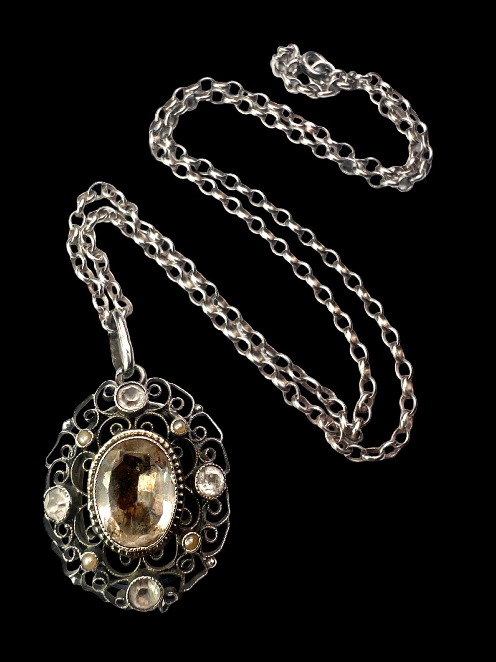 An antique silver filigree oval locket pendant featuring a central oval faceted stone surrounded by delicate wirework scrollwork and seed pearl accents on a silver chain. View II