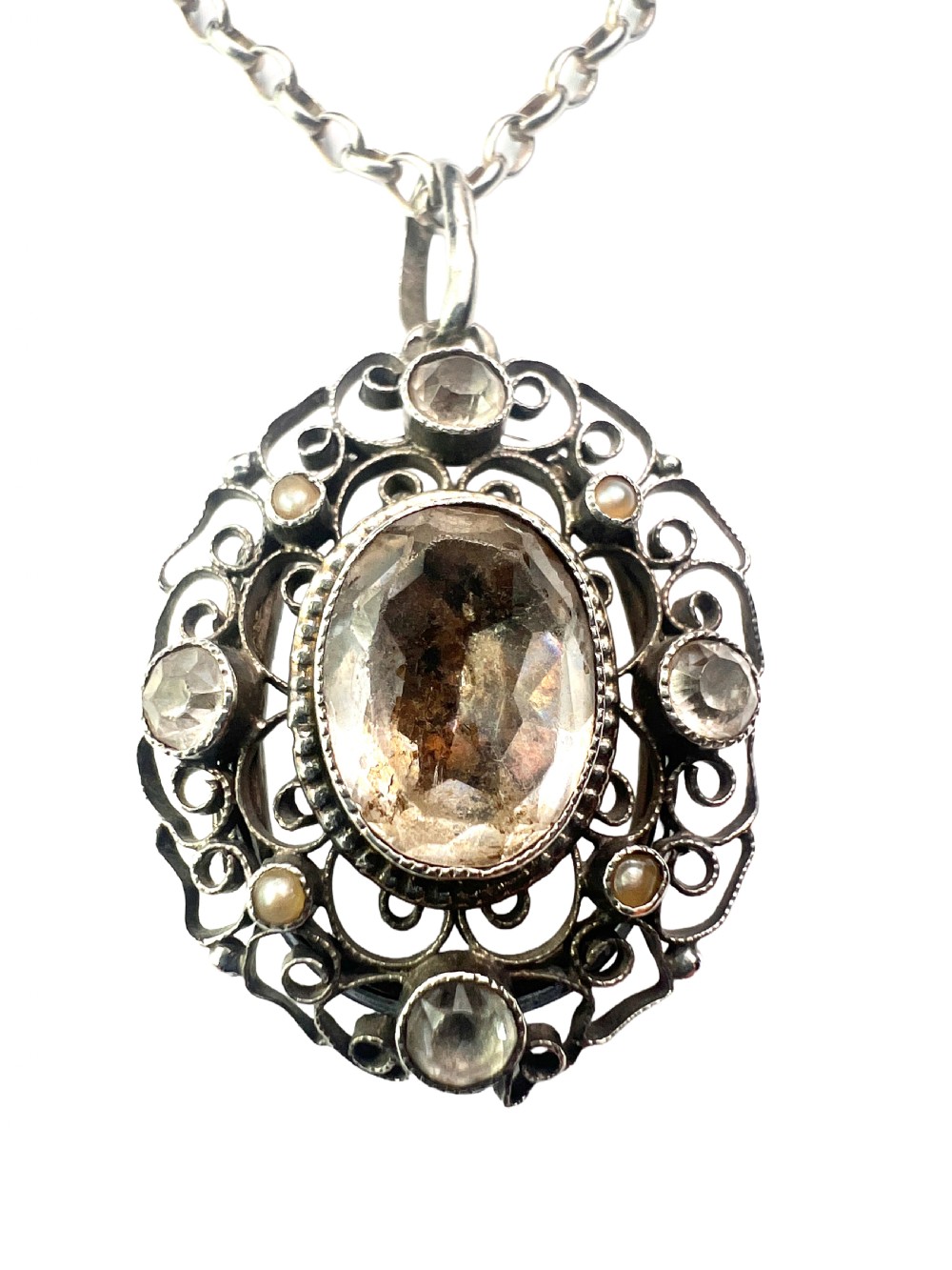 An antique silver filigree oval locket pendant featuring a central oval faceted stone surrounded by delicate wirework scrollwork and seed pearl accents on a silver chain. View III