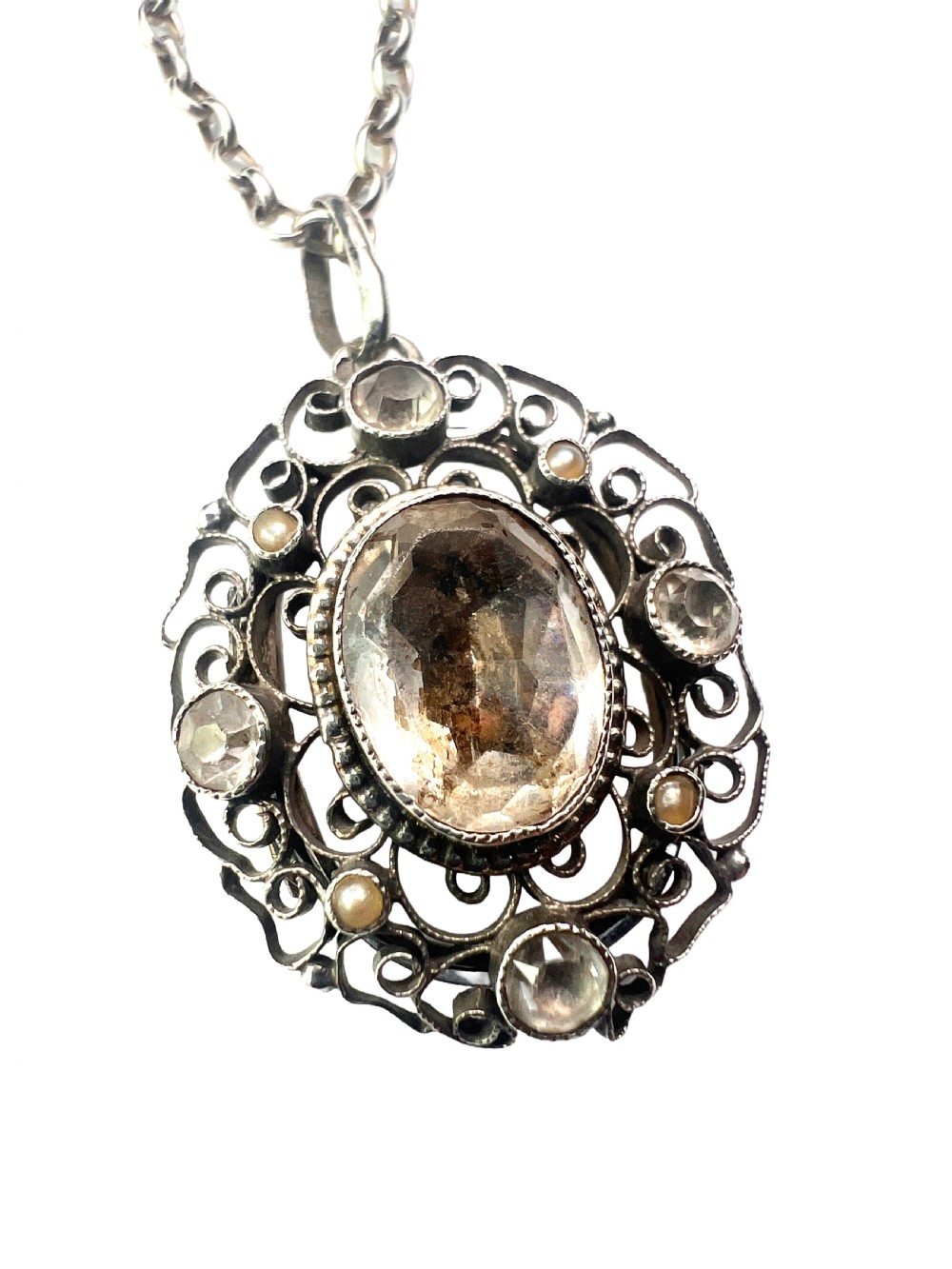 An antique silver filigree oval locket pendant featuring a central oval faceted stone surrounded by delicate wirework scrollwork and seed pearl accents on a silver chain. View VIII