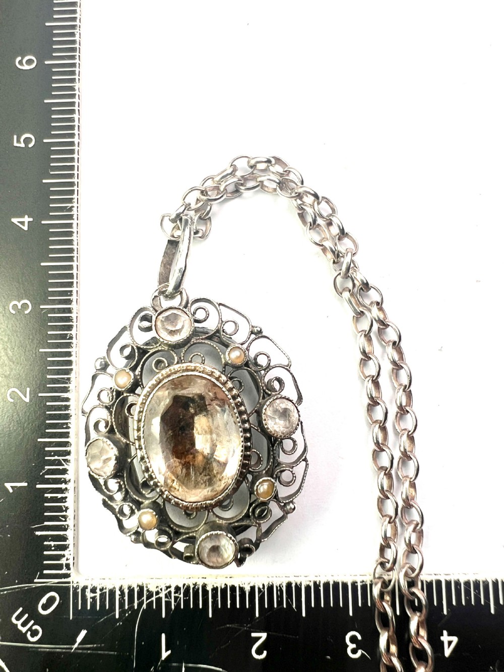 An antique silver filigree oval locket pendant featuring a central oval faceted stone surrounded by delicate wirework scrollwork and seed pearl accents on a silver chain. View IX