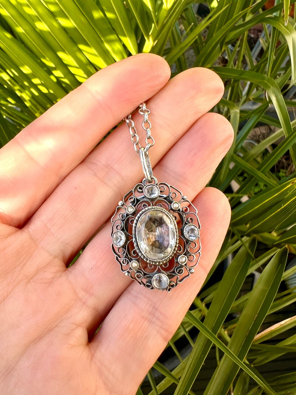 An antique silver filigree oval locket pendant featuring a central oval faceted stone surrounded by delicate wirework scrollwork and seed pearl accents on a silver chain. View X