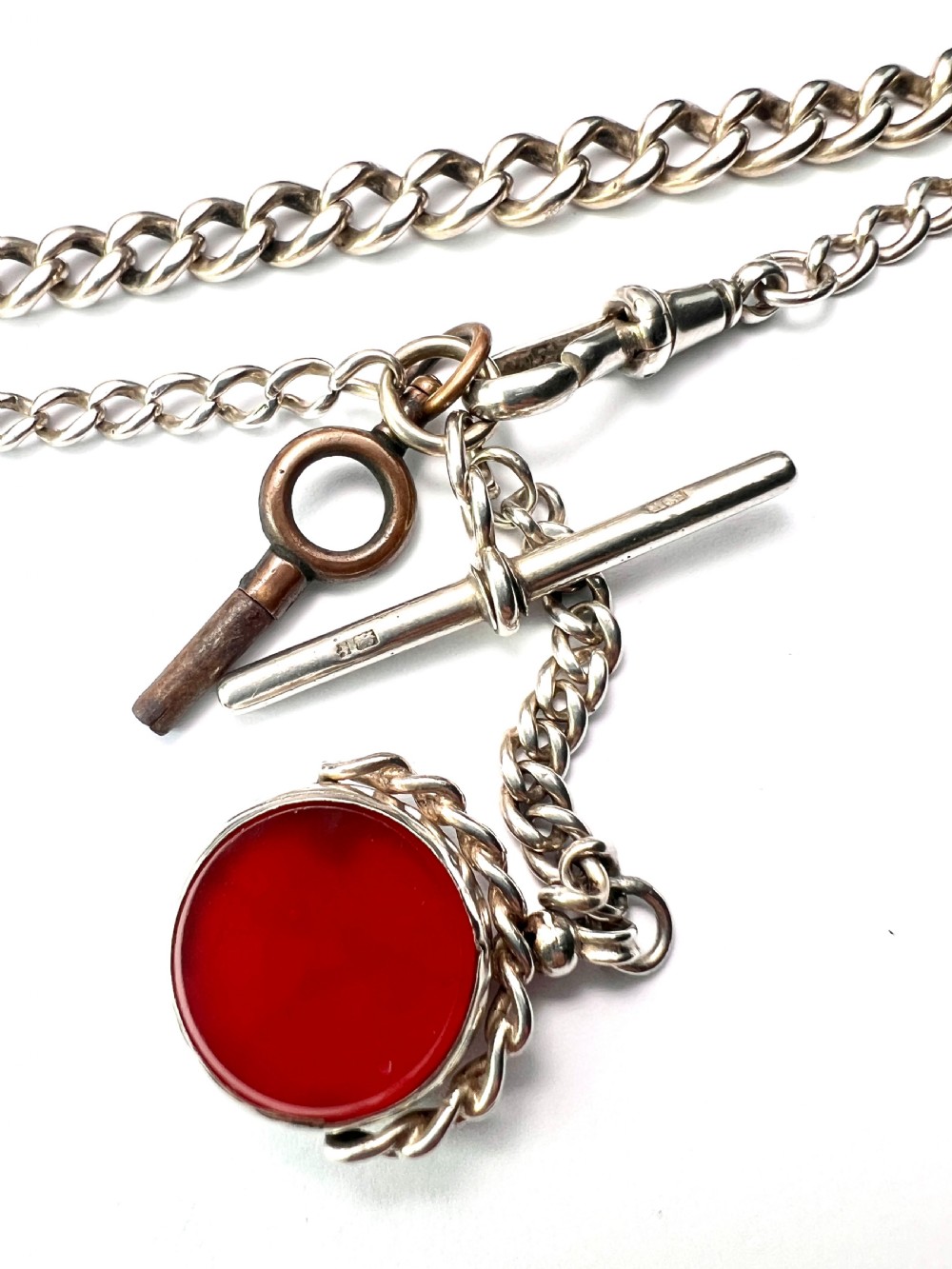 A Victorian sterling silver curb link Albert watch chain featuring a T-bar, watch key charm, and red stone swivel fob. View VI
