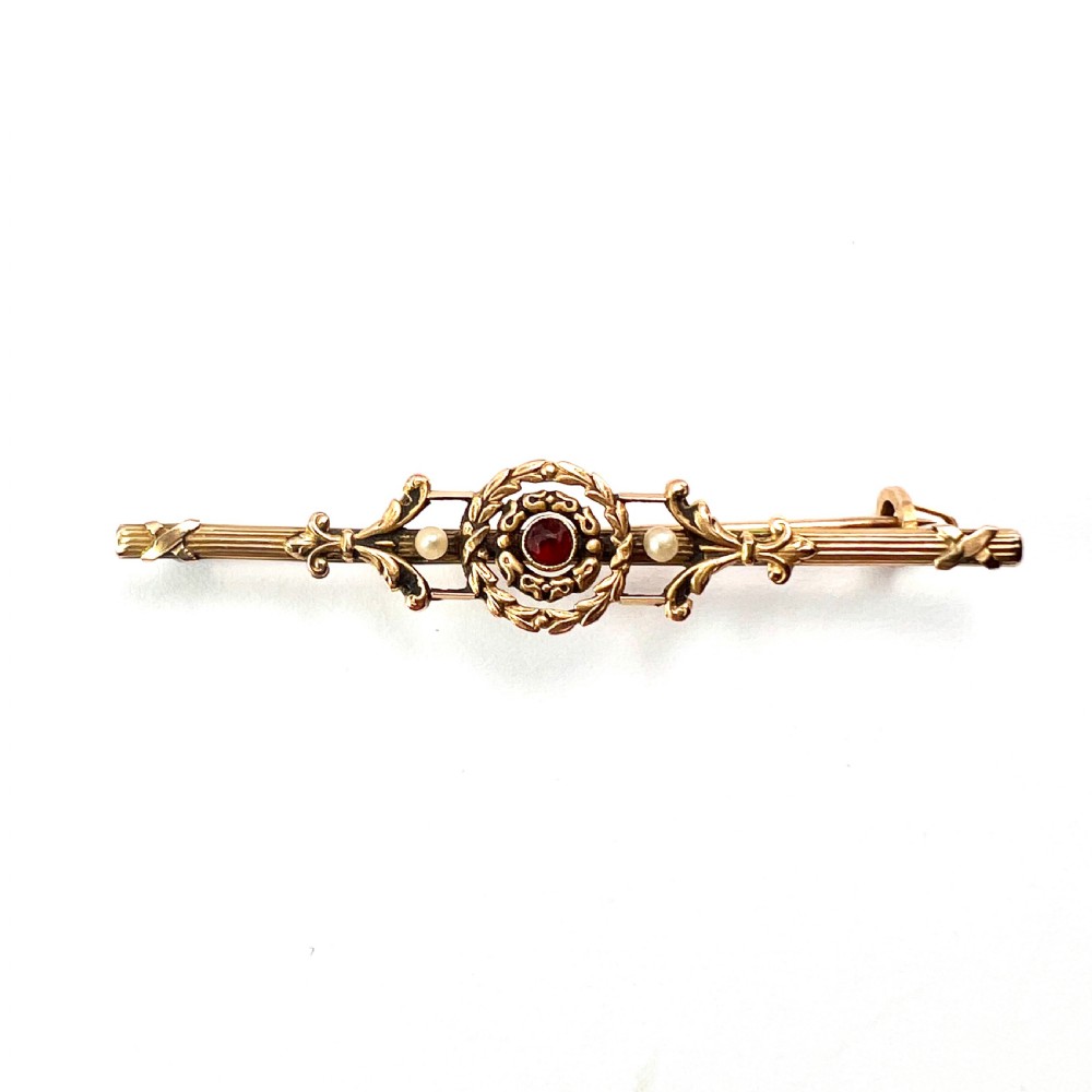 An antique 18-carat yellow gold bar brooch featuring a central circular motif with a round red garnet flanked by seed pearls and fluted bar details.