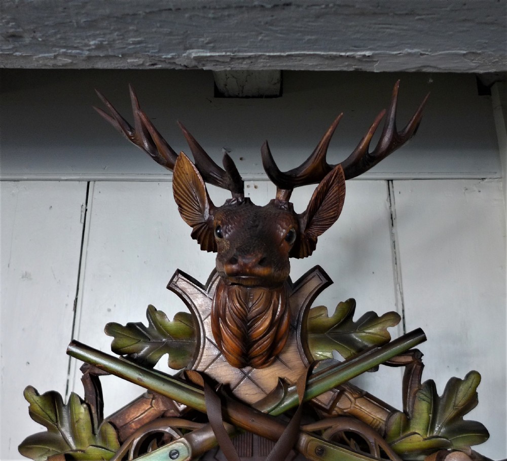 A hand-carved dark wood Black Forest cuckoo clock featuring a stag head crest, crossed rifles, game animals, and oak leaves around a central dial with Roman numerals. View II