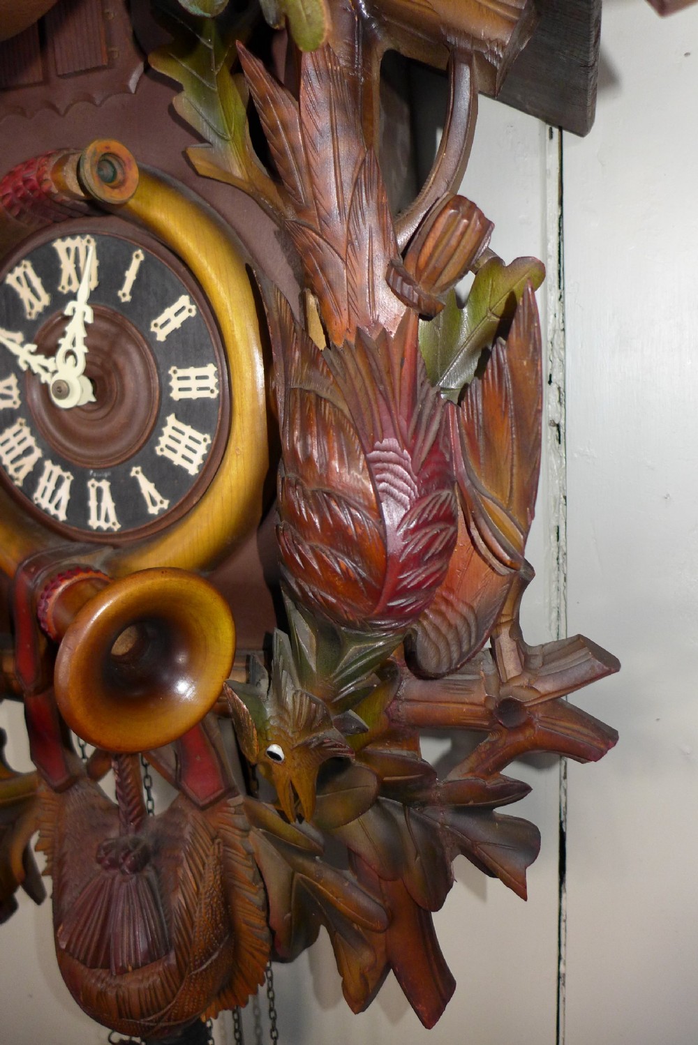 A hand-carved dark wood Black Forest cuckoo clock featuring a stag head crest, crossed rifles, game animals, and oak leaves around a central dial with Roman numerals. View V
