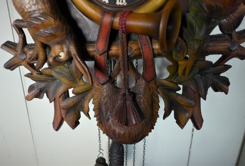 A hand-carved dark wood Black Forest cuckoo clock featuring a stag head crest, crossed rifles, game animals, and oak leaves around a central dial with Roman numerals. View VI