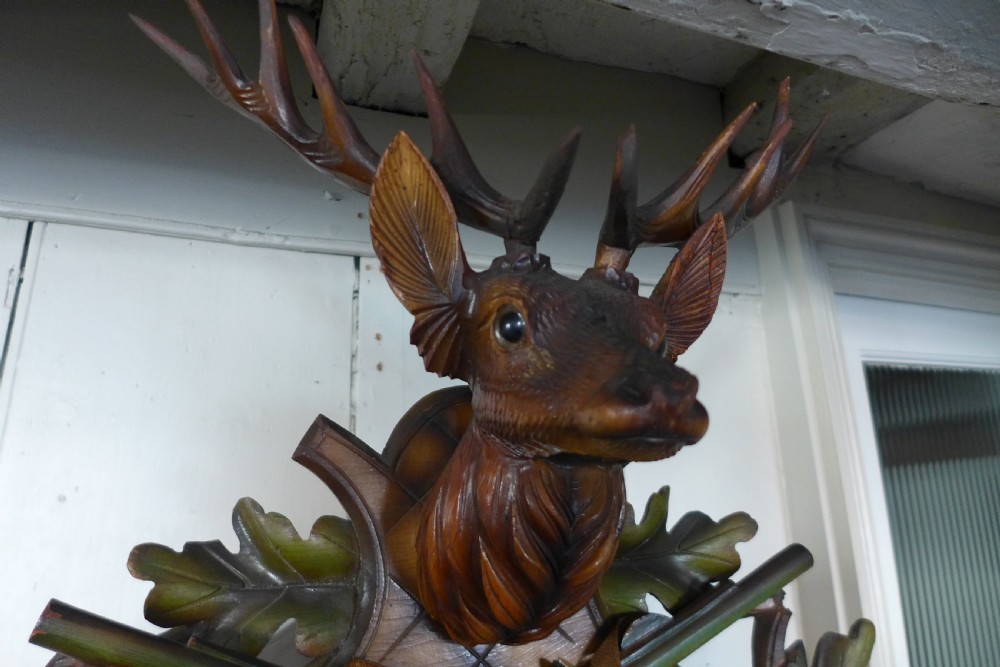 A hand-carved dark wood Black Forest cuckoo clock featuring a stag head crest, crossed rifles, game animals, and oak leaves around a central dial with Roman numerals. View VII