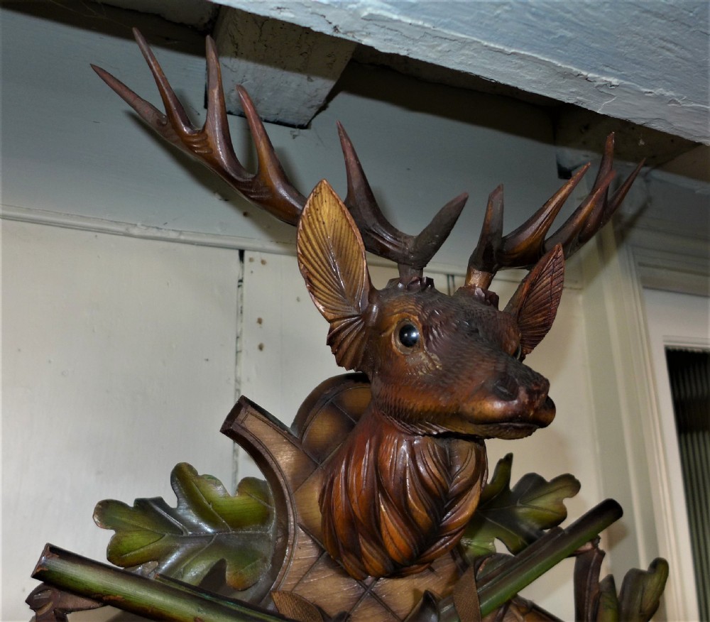 A hand-carved dark wood Black Forest cuckoo clock featuring a stag head crest, crossed rifles, game animals, and oak leaves around a central dial with Roman numerals. View VIII
