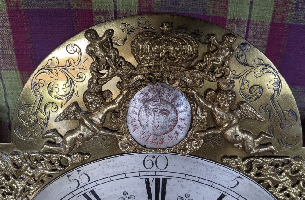 An early 18th-century brass longcase clock dial featuring an arched top with cherub spandrels, a central engraved sun face, and a silvered chapter ring with Roman numerals signed W. Reynolds Uxbridge. View V