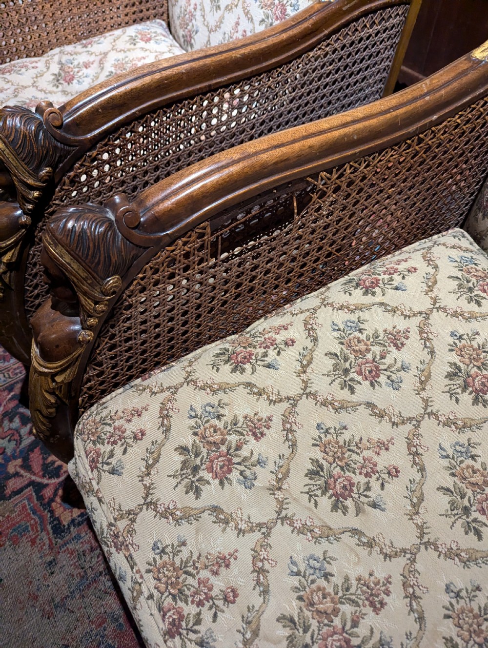 A pair of carved wood bergere armchairs featuring floral pattern upholstered cushions, caned outer arms, caryatid armrests with parcel gilding, and carved paw feet. View V