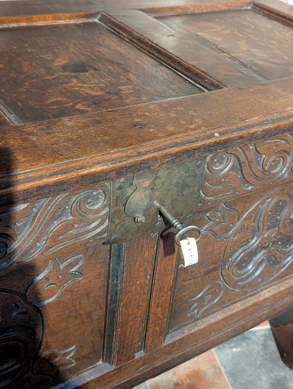 A small 17th-century dark oak coffer featuring carved foliate front panels, a three-panel hinged lid, and an iron lock plate. View V