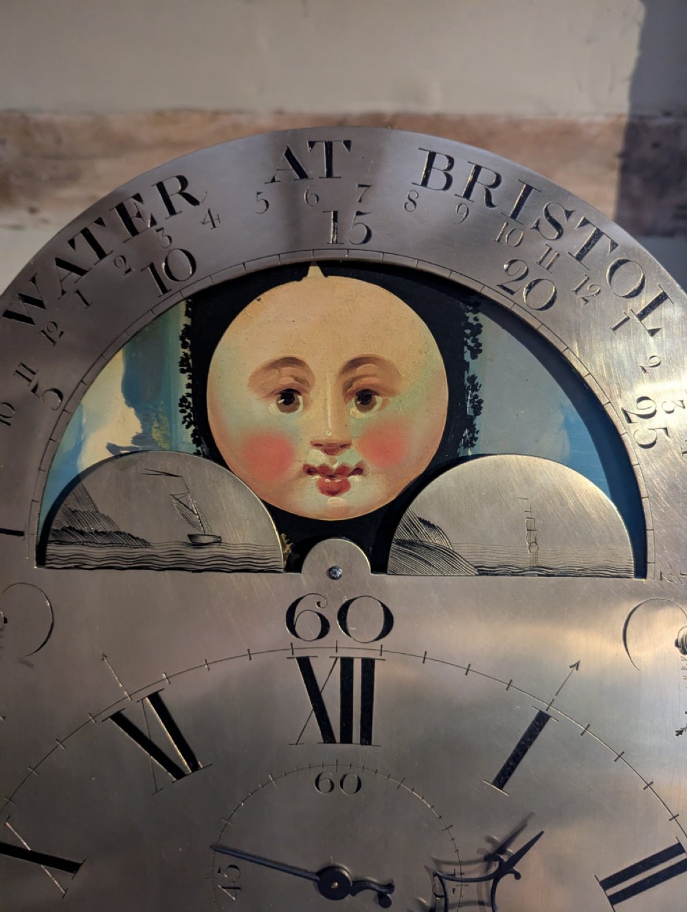 An early nineteenth-century flame mahogany longcase clock featuring a swan-neck pediment, brass dial with a painted moon phase arch, and oval stringing on the trunk door. View V