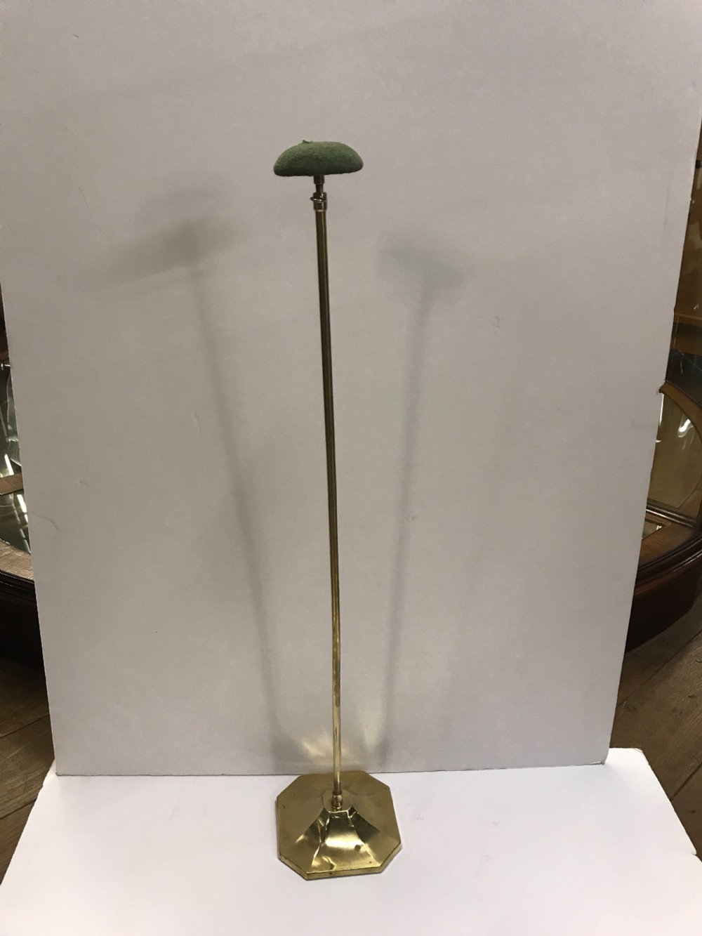 A tall antique brass shop hat display stand featuring a faceted base, a slender column, and a green fabric cushioned top.