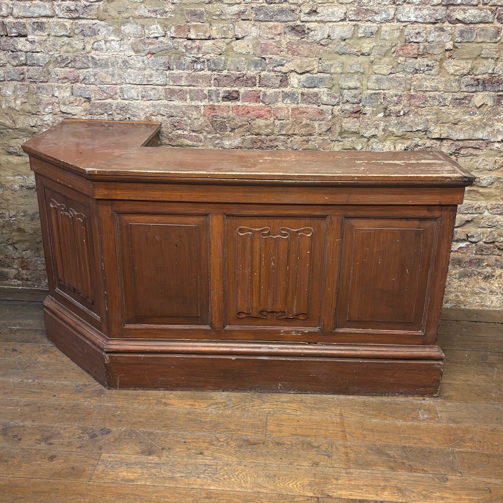 An angled Edwardian oak commercial shop counter featuring recessed timber panels with carved decorative motifs and a heavy plinth base.