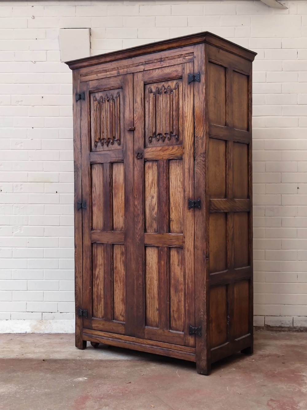 A tall solid oak double wardrobe featuring carved linenfold upper panels and black iron strap hinges.