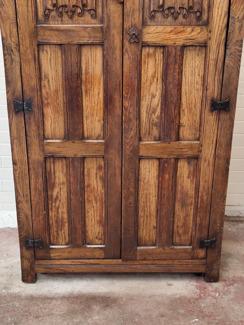 A tall solid oak double wardrobe featuring carved linenfold upper panels and black iron strap hinges. View XII